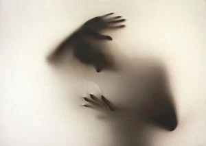 eine Schattenfigur, umgeben von warmem Licht; den Kopf gesenkt; ene Hand - wie abwehrend - über dem Kopf, die andere vor der Brust